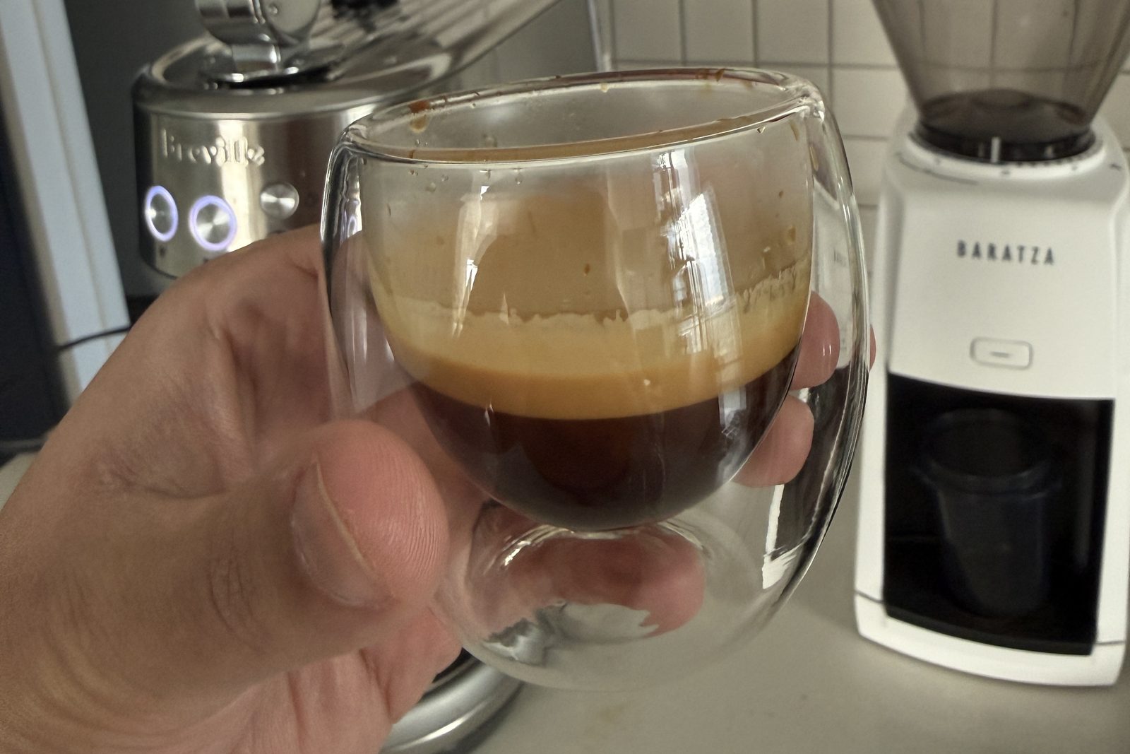 A shot of espresso in a double walled glass beside a Breville Bambino and a Baratza Encore ESP